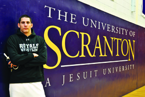 Male student standing in front of "The ɫtv  A Jesuit University" sign on wall.